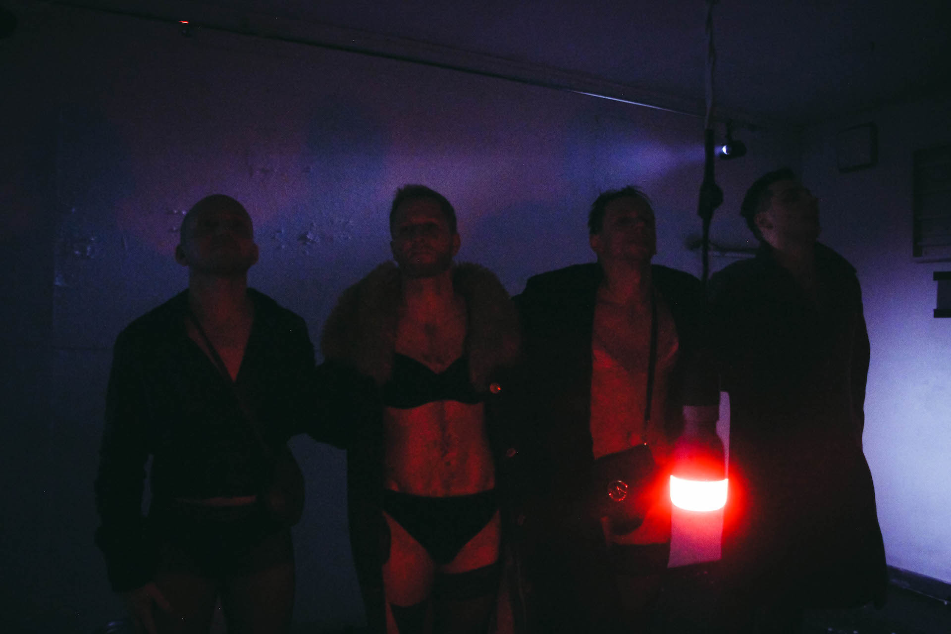 In a dark blue lighted room, a red light bulb is hanging by the ceiling. Four performers are standing behind the light bulb in one line. They are all wearing underwear and long jackets