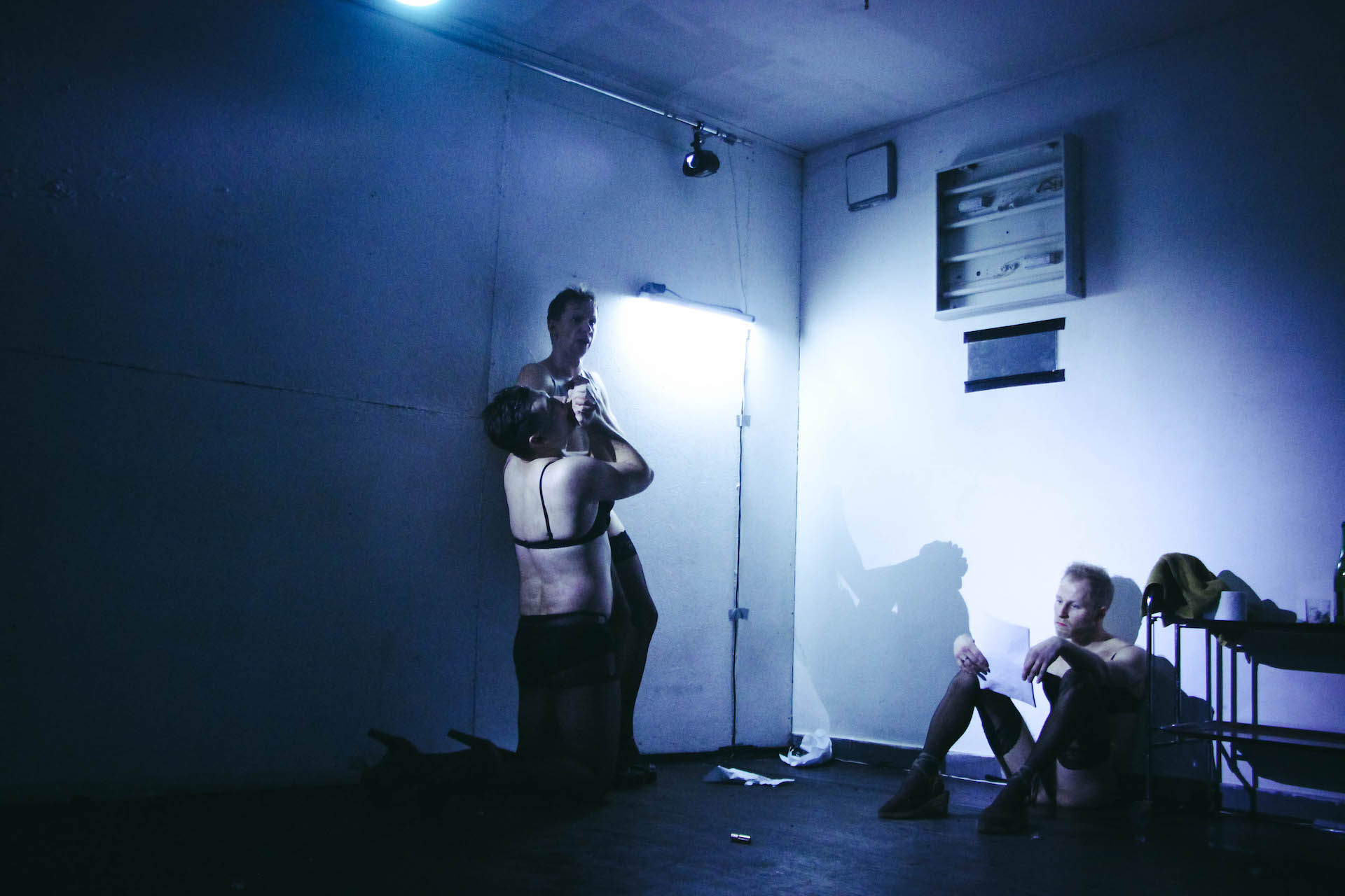 Three performers in a dark blue lighted room. They are wearing a bra, pantyhose and high heels. One on the right is sitting on the floor leaned against the wall, reading something from piece of paper. Two performers are on the left. One is kneeling down and kissing the hand of the other one standing, as if they are praying.