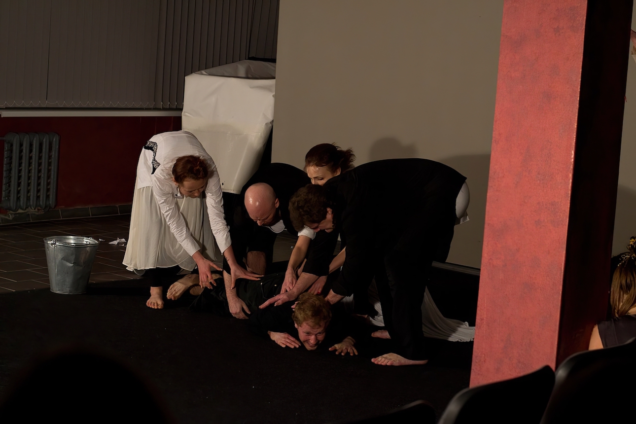 Four performers in black and white are pushing one performer in black shirt and trouser to the floor.