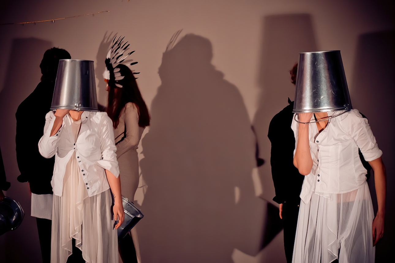Two performers wearing a white chiffon dress and a white blouse is standing with a metal bucket over their heads.
