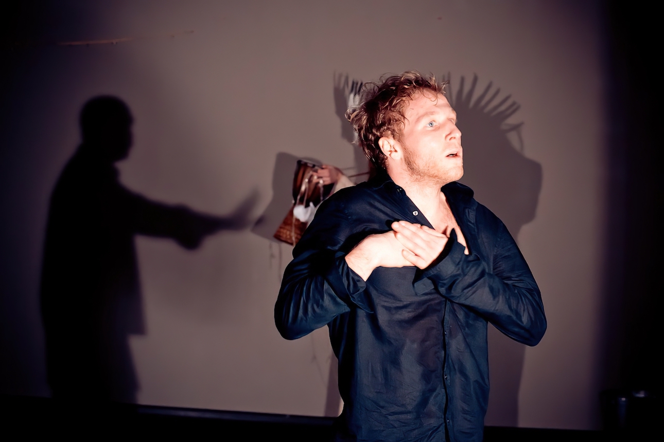 One performer wearing a black shirt is looking at the front with both of their hands on the right chest. Behind, there is a shadow of a performer interacting with another performer with a headdress and djembe