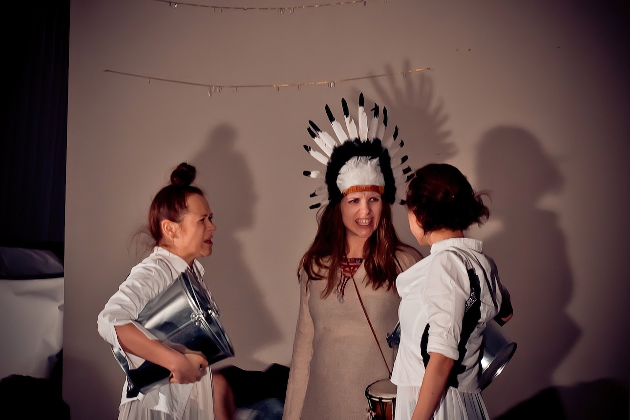 Two performers with a white chiffon dress and a white blouse are each holding a metal bucket. They are arguing with another performer in beige dress and headdress.