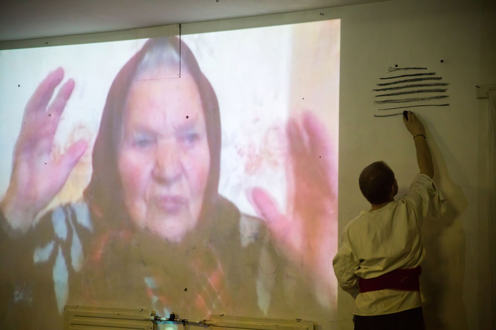 A video of an old lady is projected on the wall. Meanwhile, a performer in a white shirt is drawing black lines with black chalk next to the projected wall.