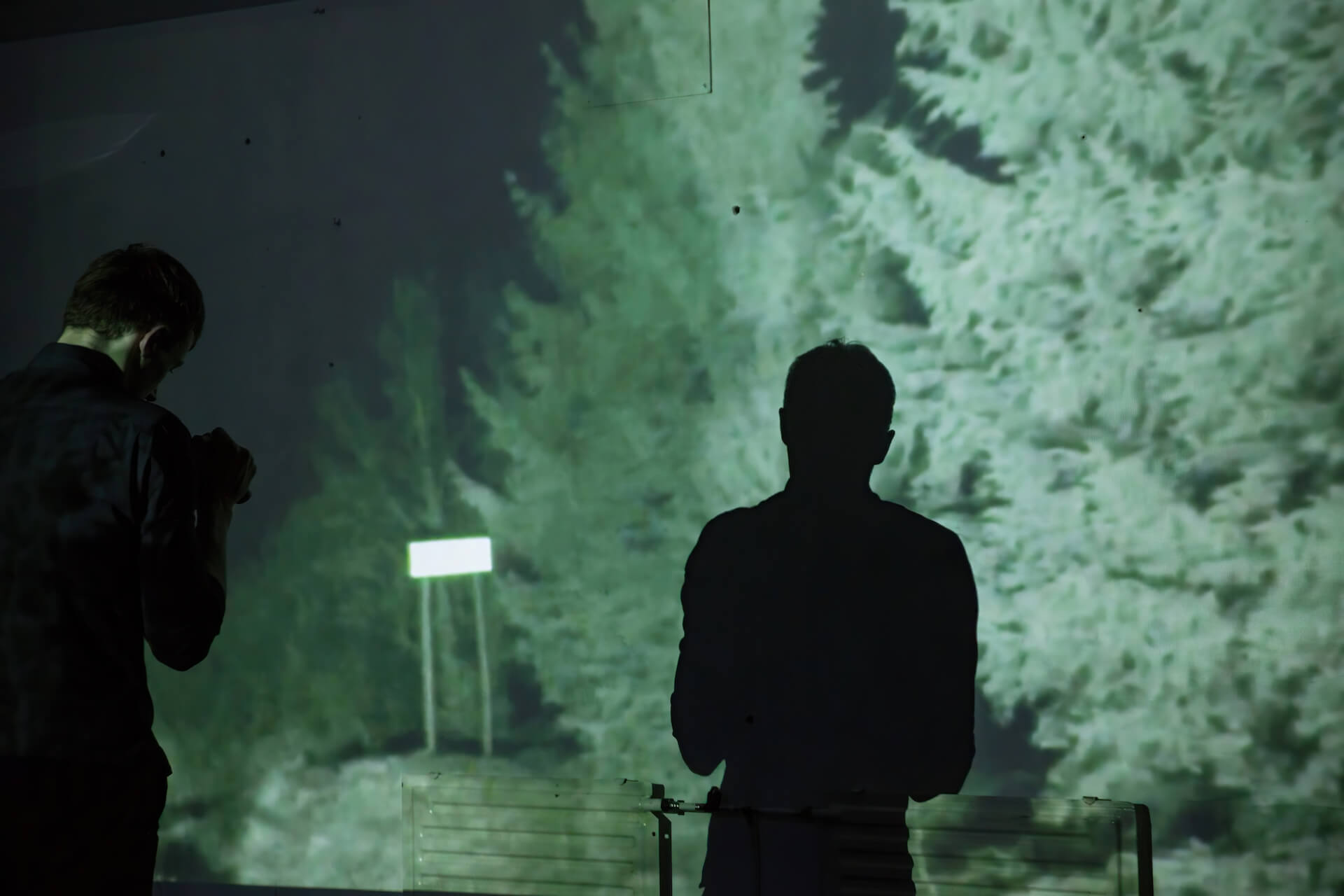 In the background, a picture of a dark forest is projected on the wall. In front of it stands two performers facing towards the wall. The room is so dark that there is only the silhouette of the two performers and the projected image on the wall.