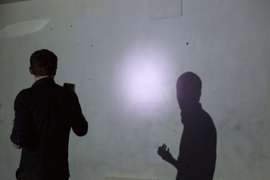 A performer stands in front of a white wall shining a flashlight on the wall. There is the performer and the shadow of the performer on the wall.