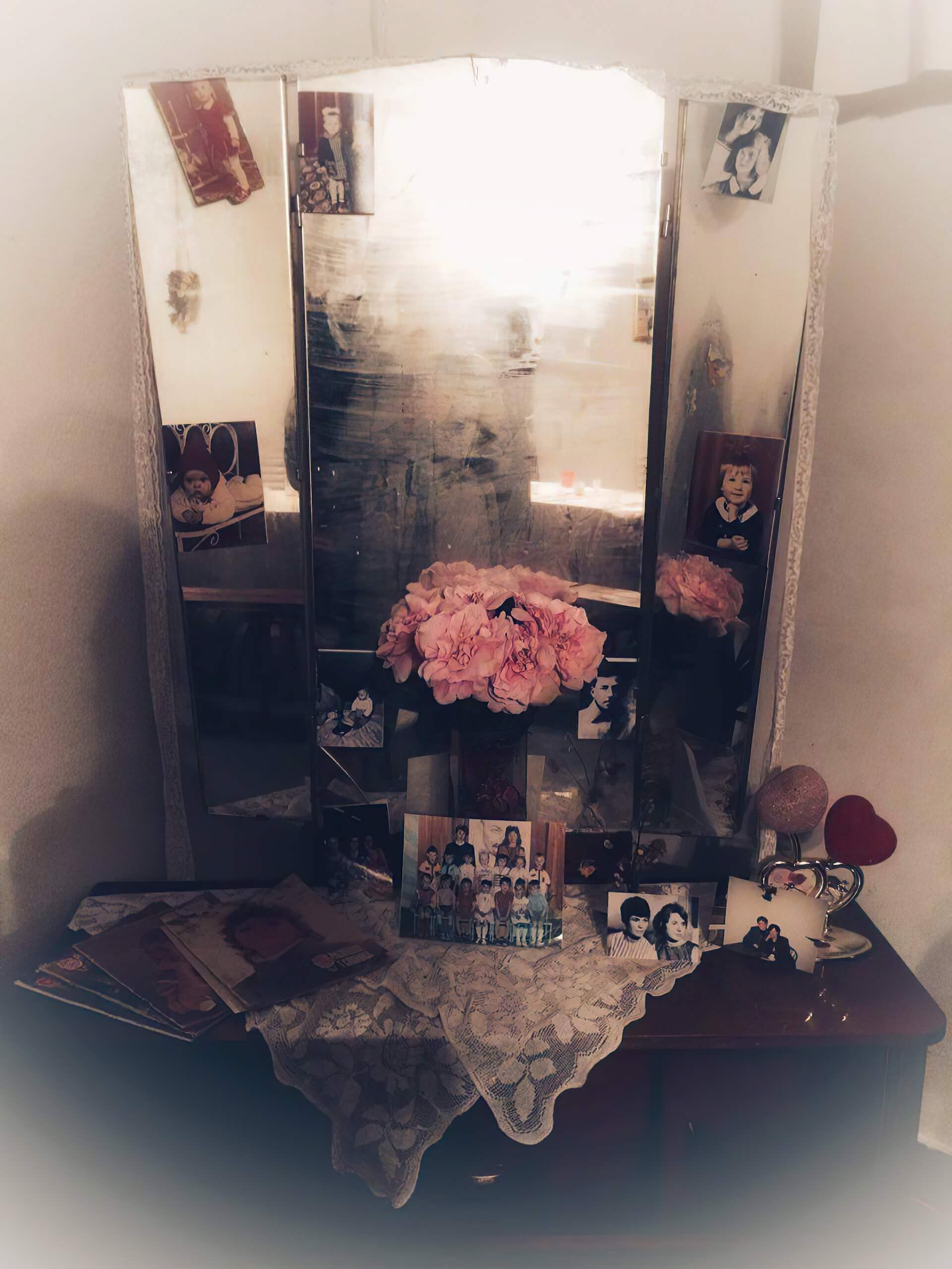 Three sided mirror stands on top of a dressing table. There are some photos of young children on the mirror. There is a glass vase with pink roses in the middle of the dressing table. The edges are blurred out to create a nostalgic atmosphere