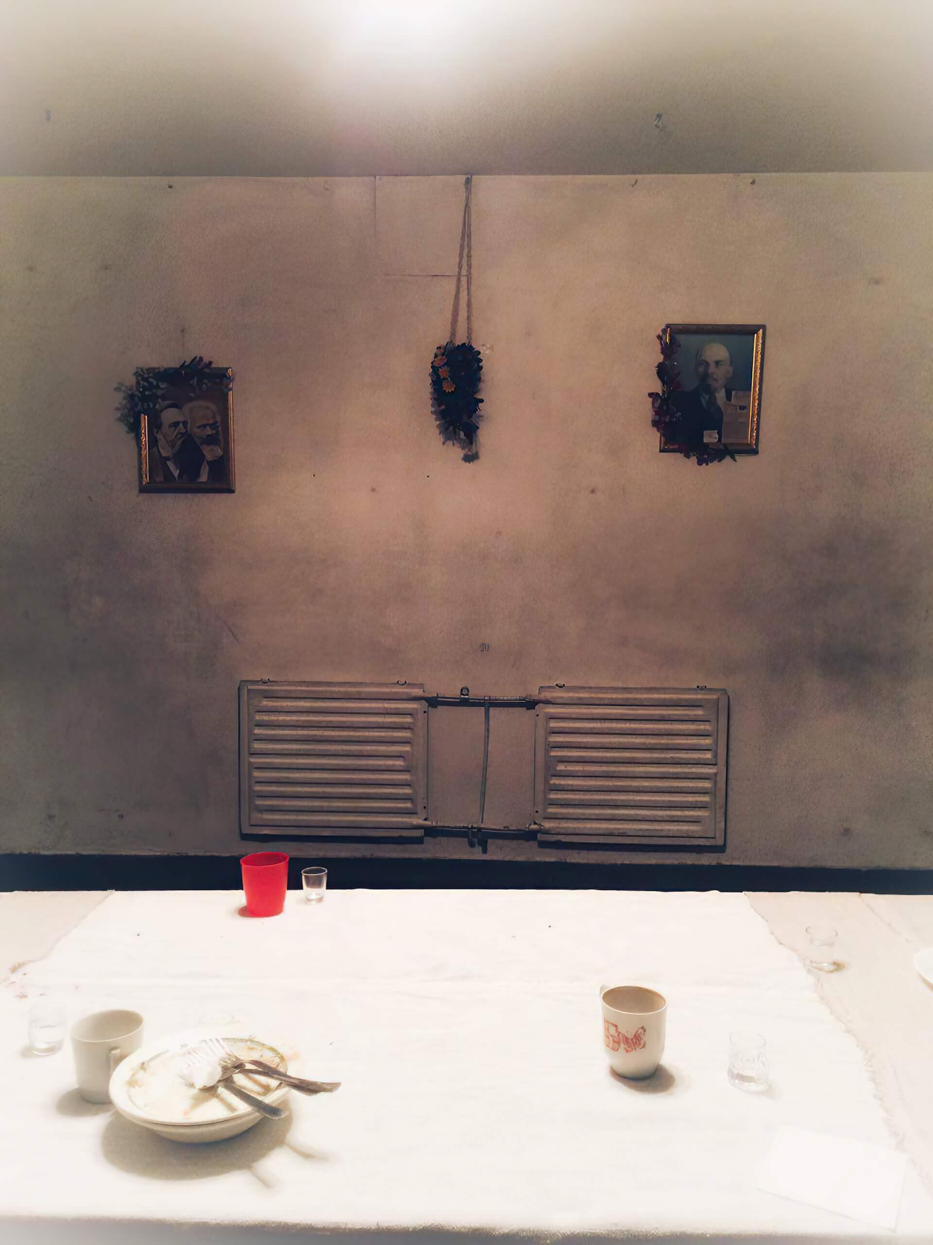 In an empty white room, there is a white table with white plates and mug cups. There is a red cup as well along with some shotglasses. Two framed pictures of someone is hanging on the wall, with the frames decorated with flower.