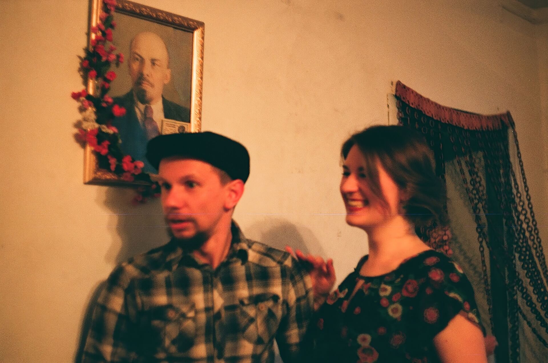 Two people standing in front of a portrait decorated with flowers. One is looking at something surprised and the other is laughing