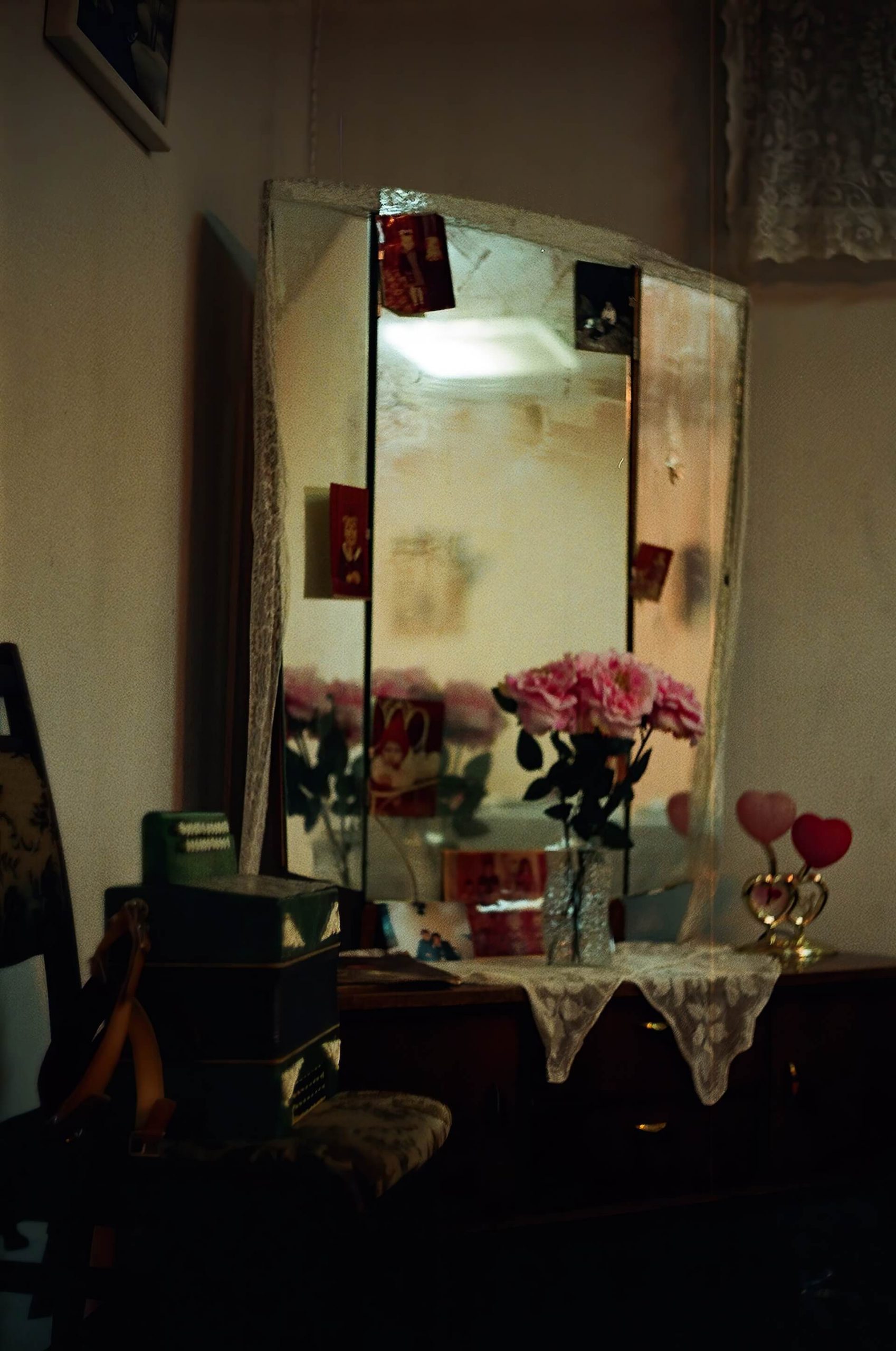 Three sided mirror stands on top of a dressing table. There are some photos of young children on the mirror. There is a glass vase with pink roses in the middle of the dressing table.