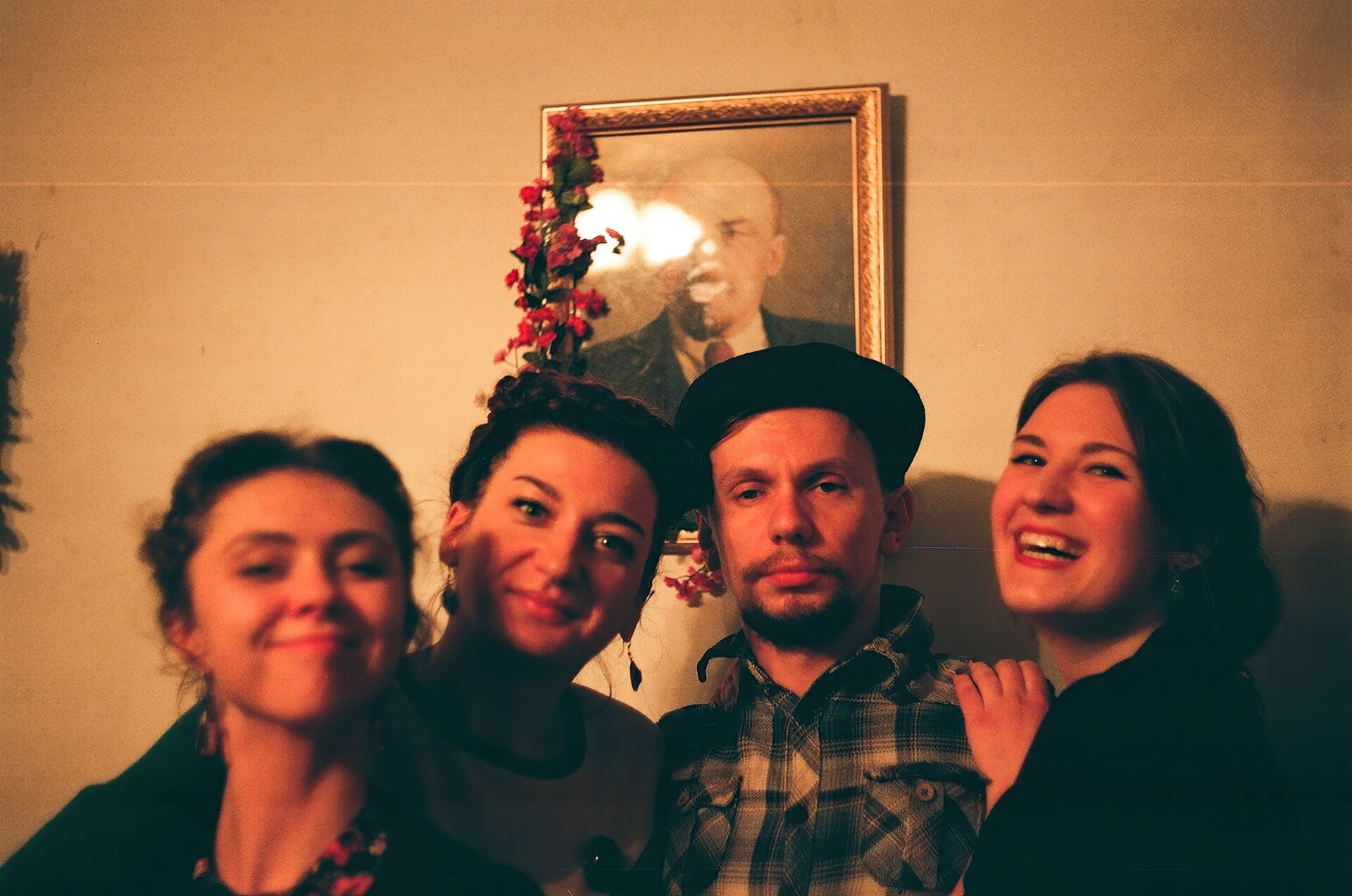 Four people are posing for photo in front of a portrait decorated with flowers. Everyone is smiling except one who looks unamused.