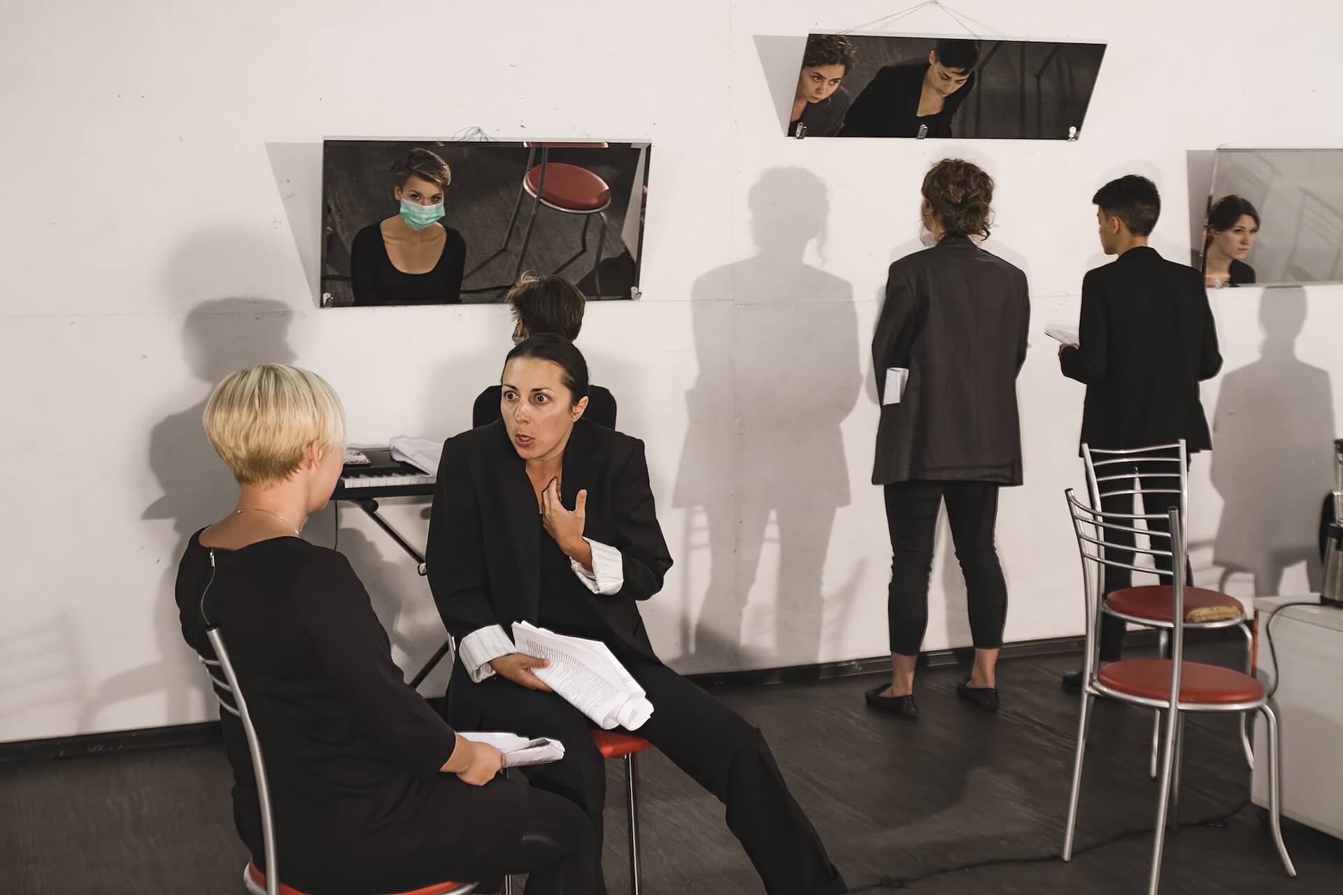 There are five performers wearing black suits in a room with three mirrors hanging by the wall. Two of them are sitting on chairs, holding a roll of script and talking to each other. One is sitting in front of one of the mirrors wearing a disposable mask. Two others are standing in front of another mirror and are talking to each other.