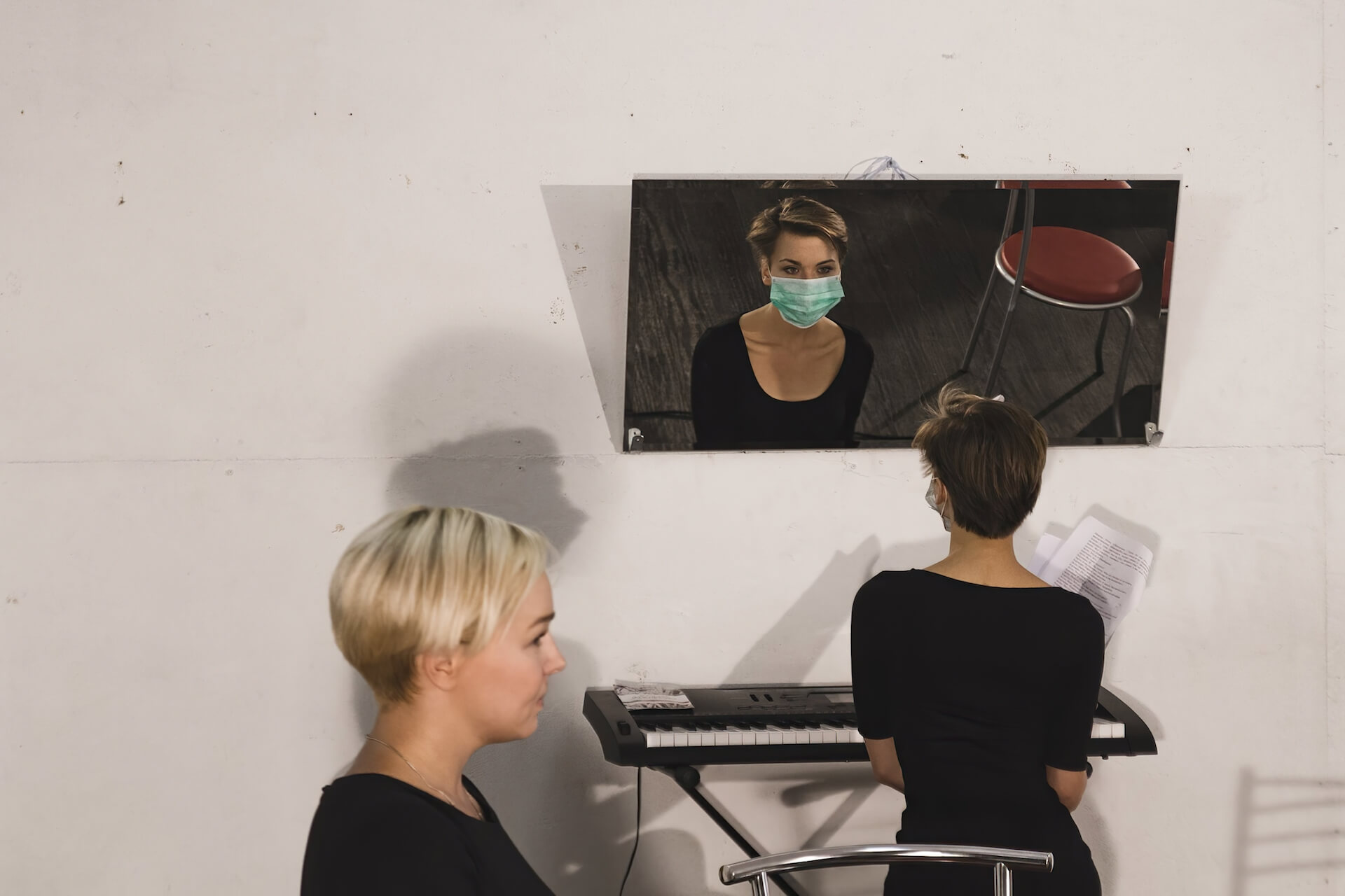 Behind shot of a performer sitting in front of a mirror wearing a disposable mask. The performer is looking at themselves. There is another performer sitting sideways behind the one with a mirror.