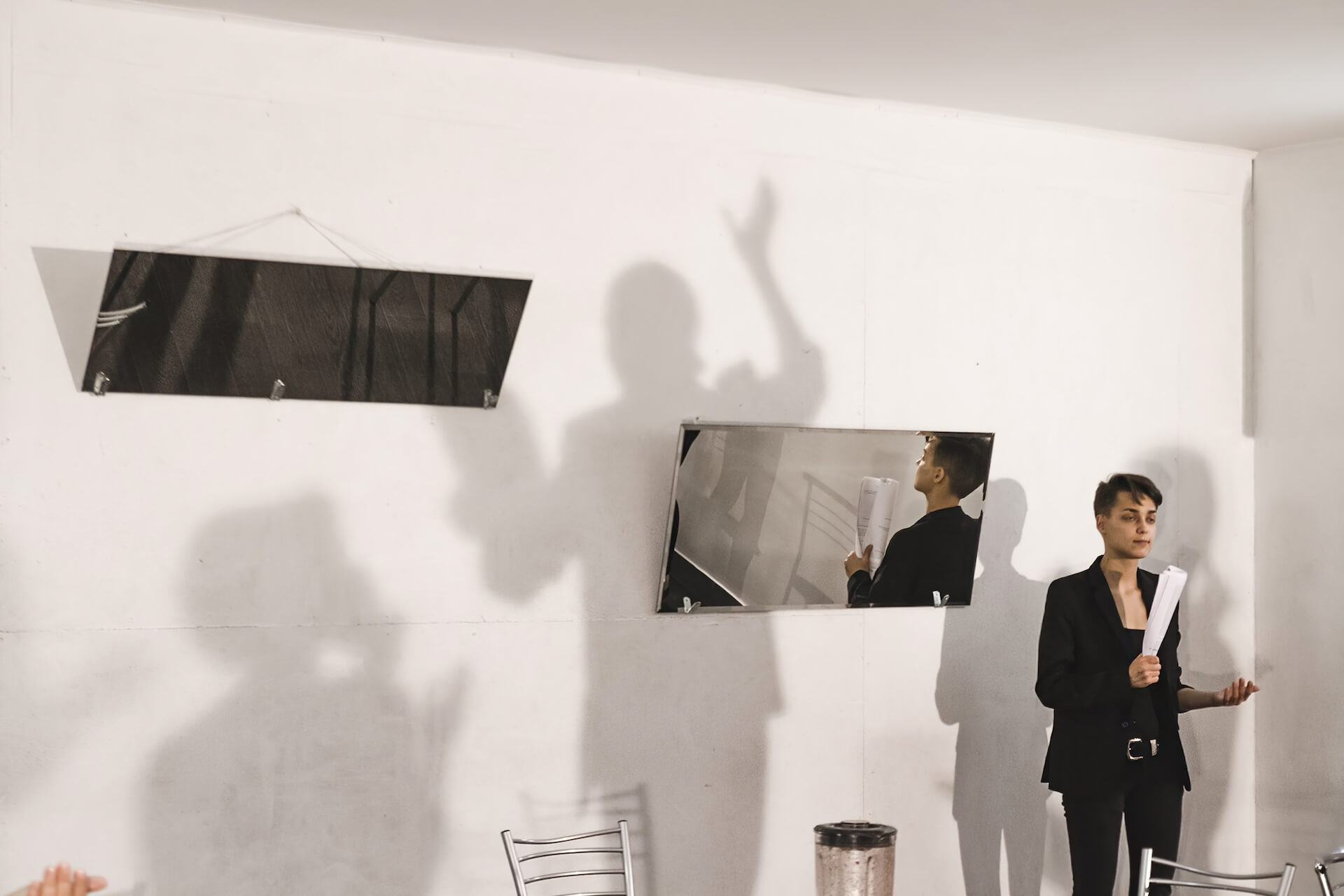 There are two mirrors hanging by the wall. On the same wall, there are shadows of performers performing with scripts on their hands. One performer stands in front of the mirror on the right, facing towards the audience.