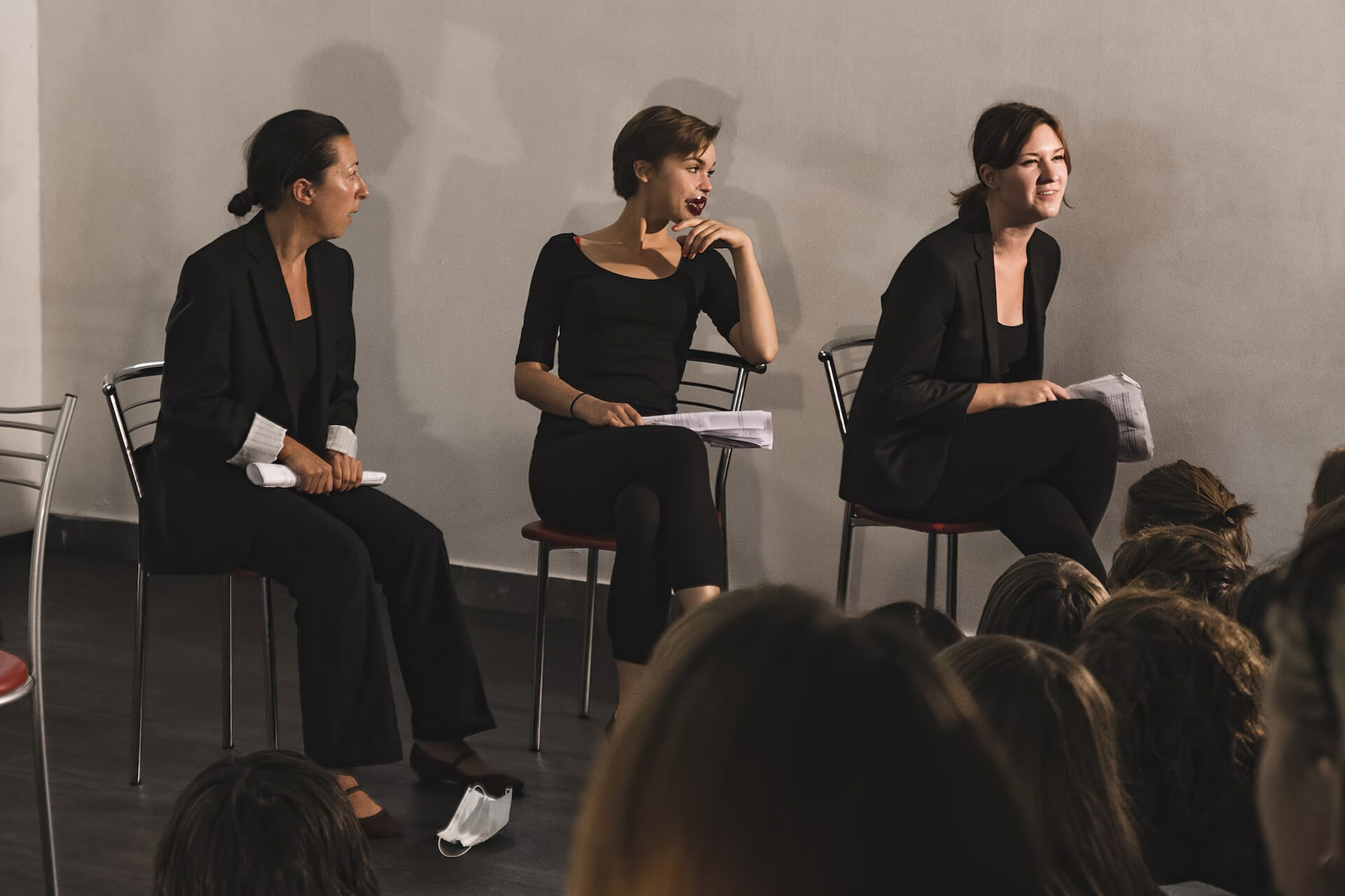 Three performers in black suits are sitting on chairs facing the audience, performing with a script in their hands. One in the middle has a large red sticker on the lips as if they recently got a massive lip filler.