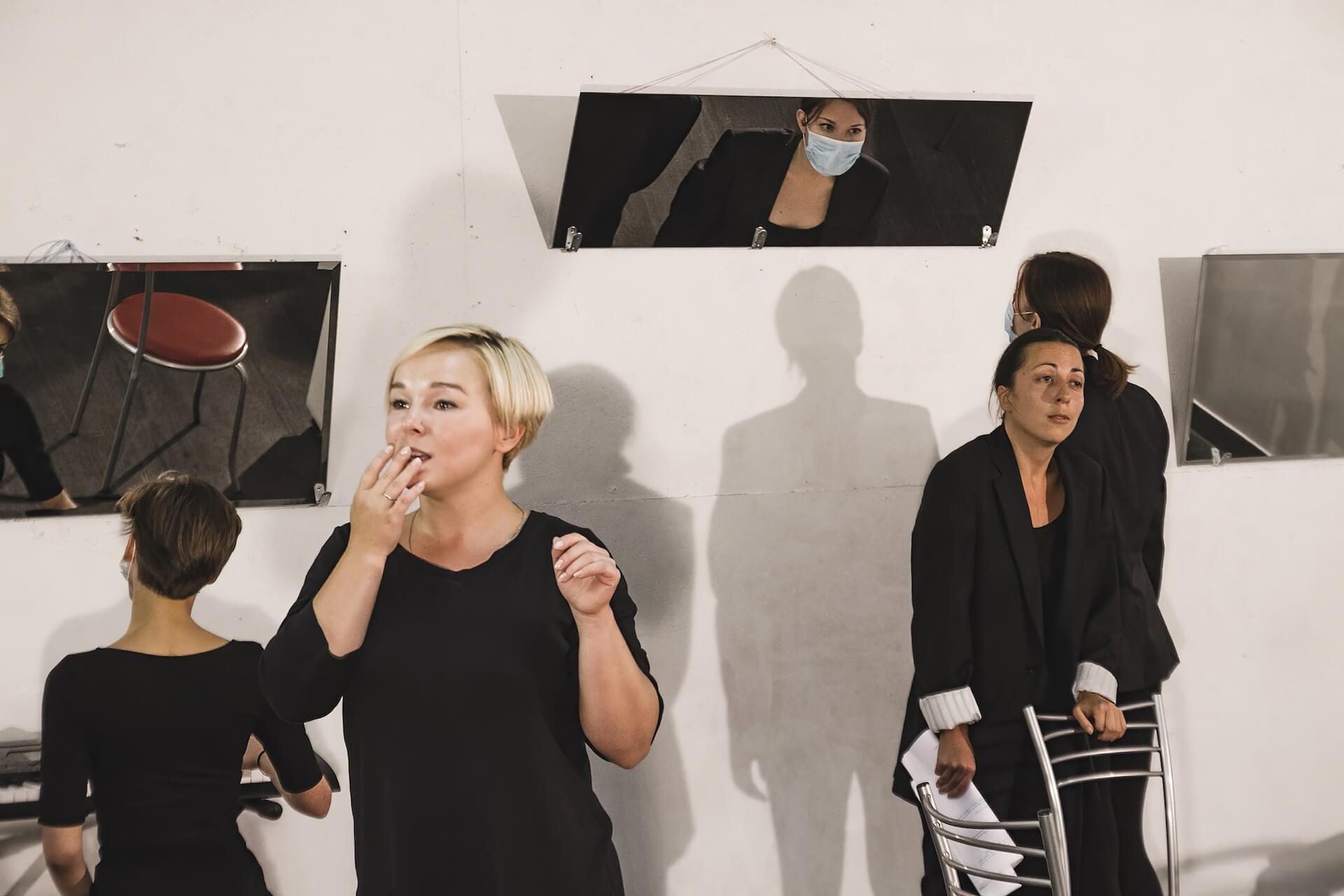 There are four performers wearing black suits in a room with three mirrors hanging by the wall. One is sitting in front of one of the mirrors wearing a disposable mask. Another one is standing in the middle wearing a disposable mask. Another one is standing behind a chair, holding a script. Another one looking at the front with their hand on the mouth.